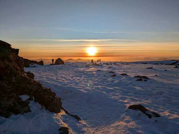 Sunset over a snowy landscape with distant silhouettes of mountains and rocks.