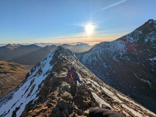 A hiker on a snowy mountain ridge, with a bright sun setting in the background.