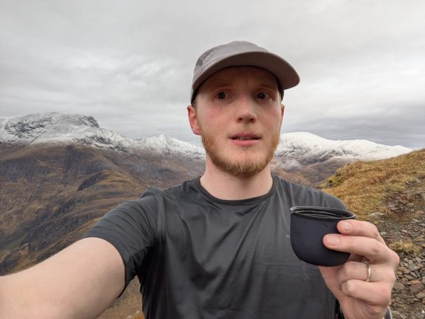 Man holding a container, standing on a mountain with snowy peaks in the background.